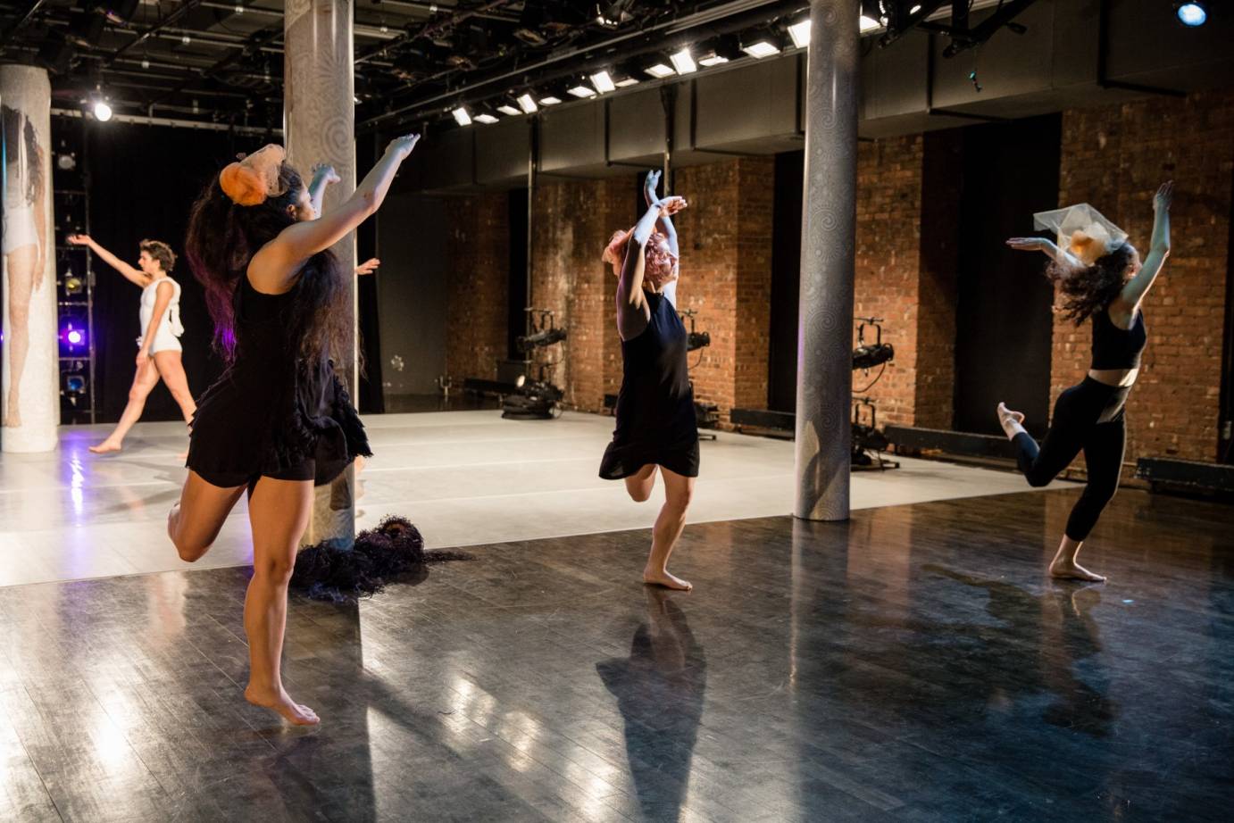 Three dancers dressed in black with orange tulle hair pieces perform on a dark wooded floor while two dancers in white dance on the other side of the stage on white marley